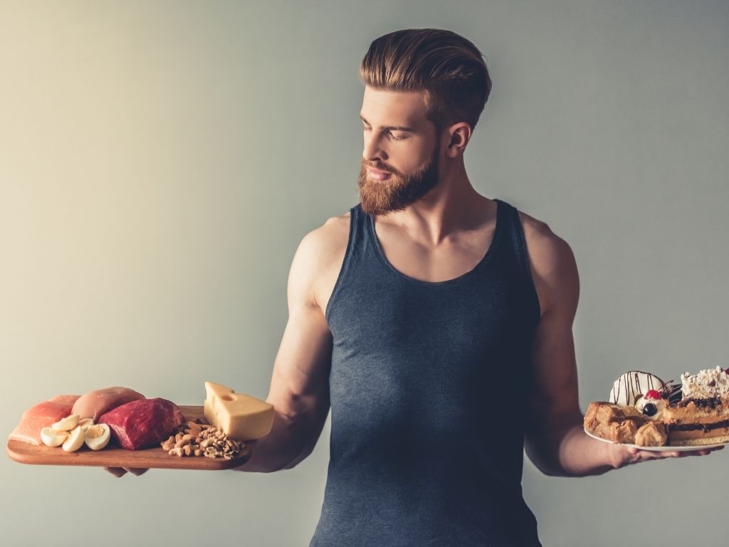young bearded sportsman holding a wooden tray with healthy food and a plate with sweets