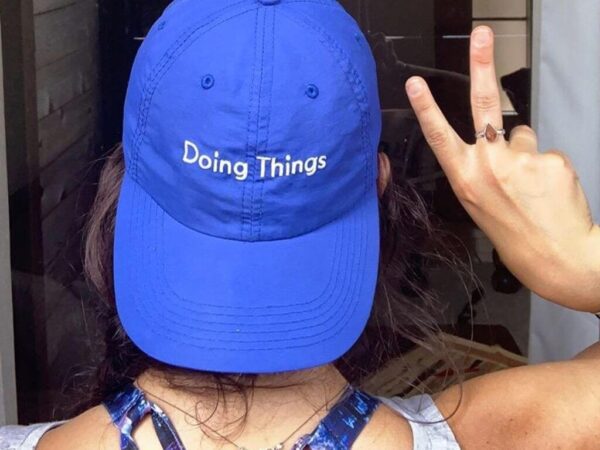 Back view of girl wearing blue hat backwards that says "Doing Things" while holding up the peace sign
