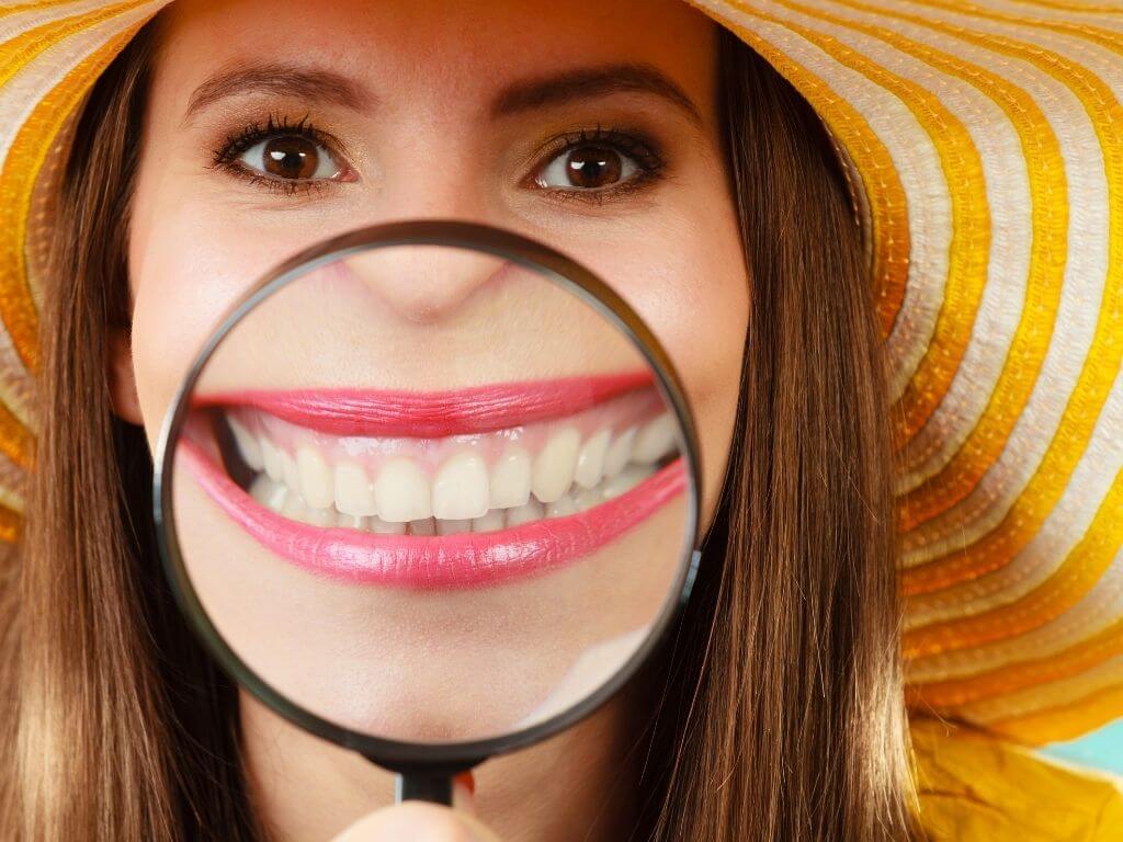 Closeup woman in summer hat smiling and showing teeth through magnifying glass